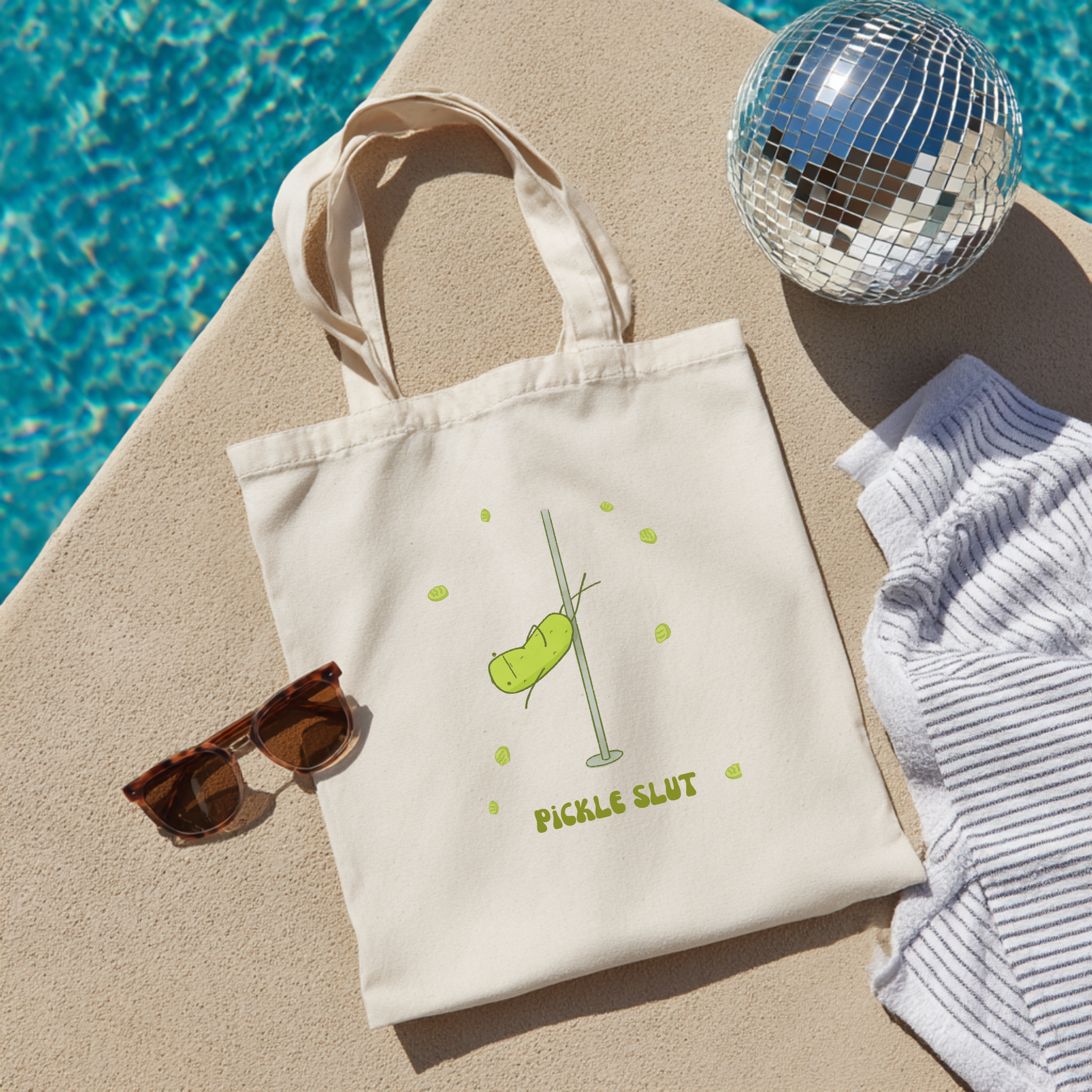 Beige tote bag with pickle design and text, sunglasses, disco ball, and striped towel on sand by pool.