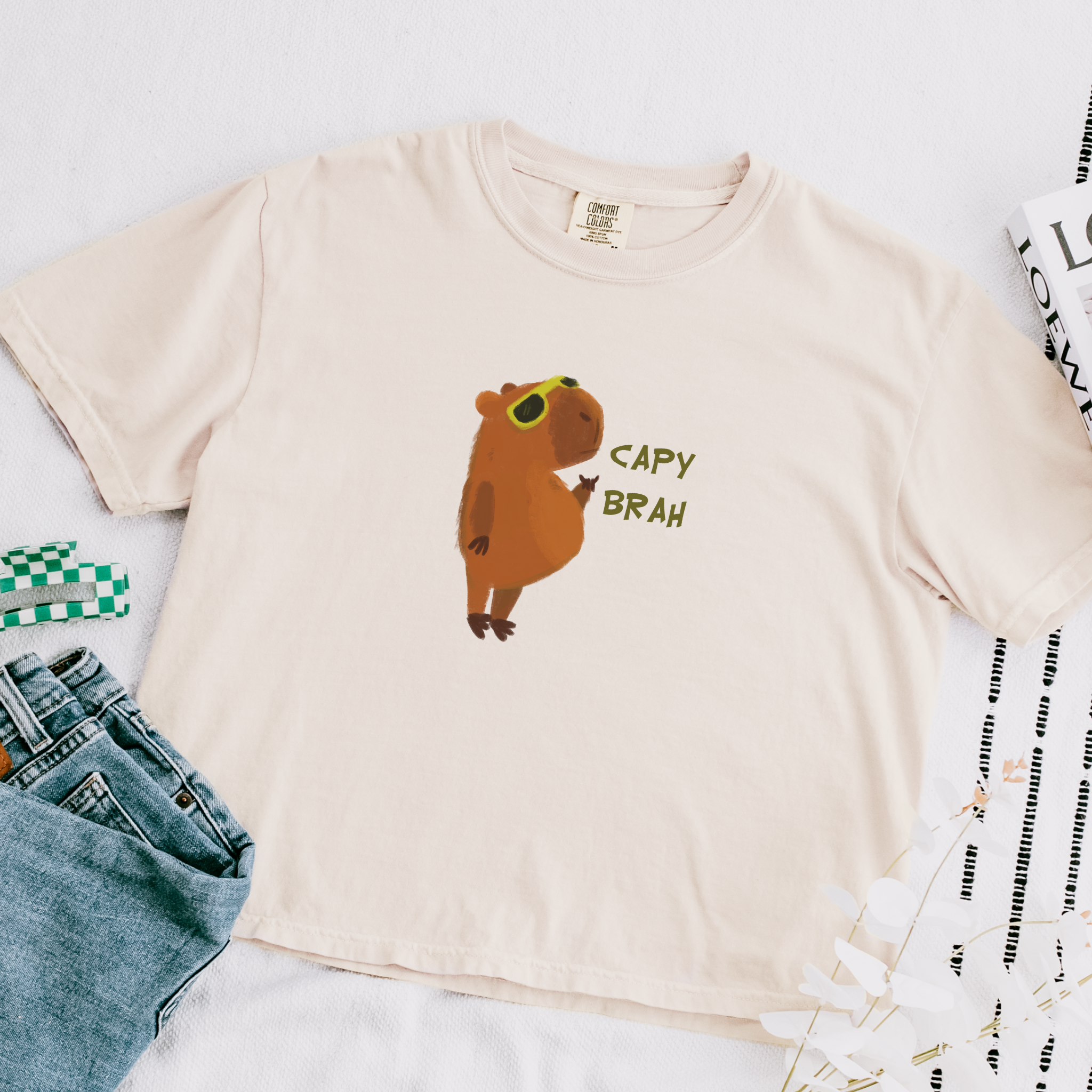 A boxy fit t-shirt featuring a graphic of a capybara with the text 'CAPY BRAH' on the front in ivory variant is laid flat with a close up view of the design