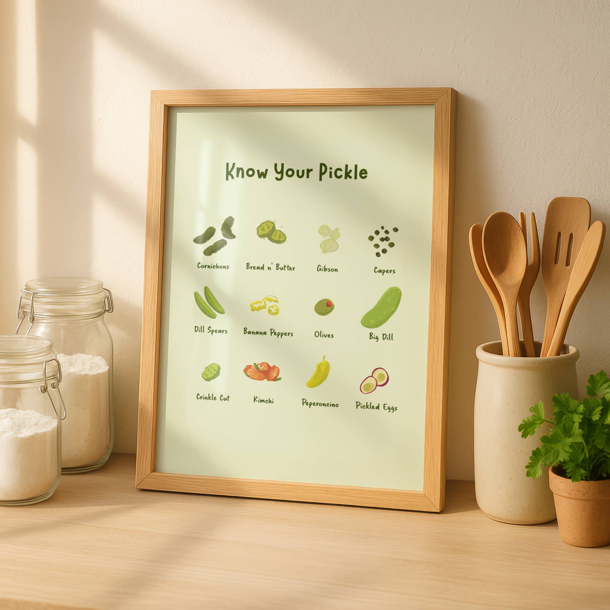 A pickle art print is displayed in a wooden frame next to some kitchen herbs and utensils