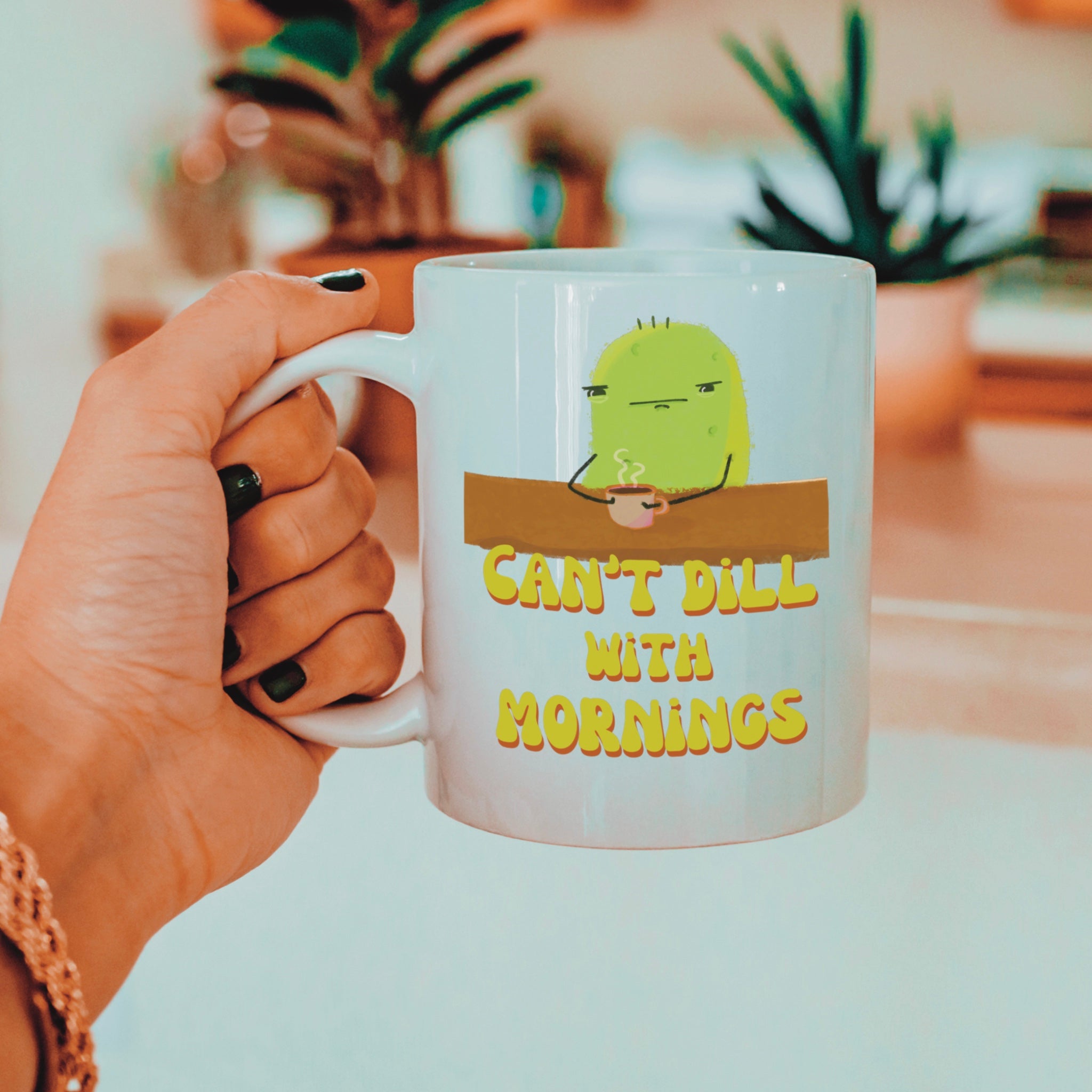 a pickle mug with a cute illustration of a pickle character drinking coffee over a punny quote that says "can't dill with mornings" is held by a woman next to house plants