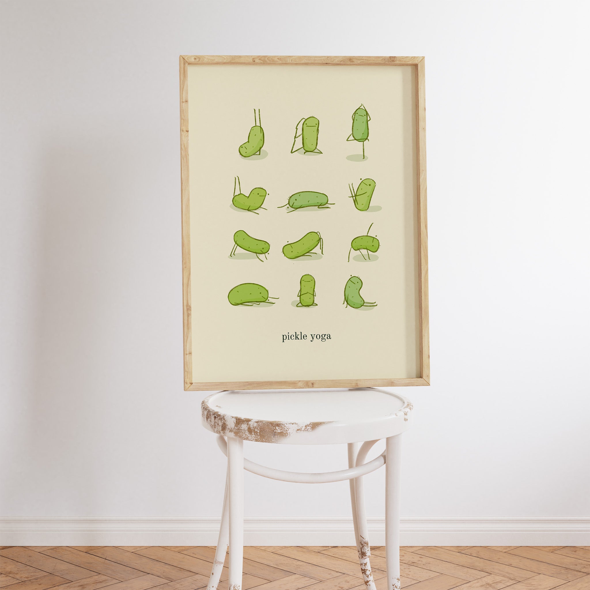 a funny pickle art print features a cute pickle in various yoga poses over text at the bottom that reads "pickle yoga" is displayed in a frame that is sitting on a white stool