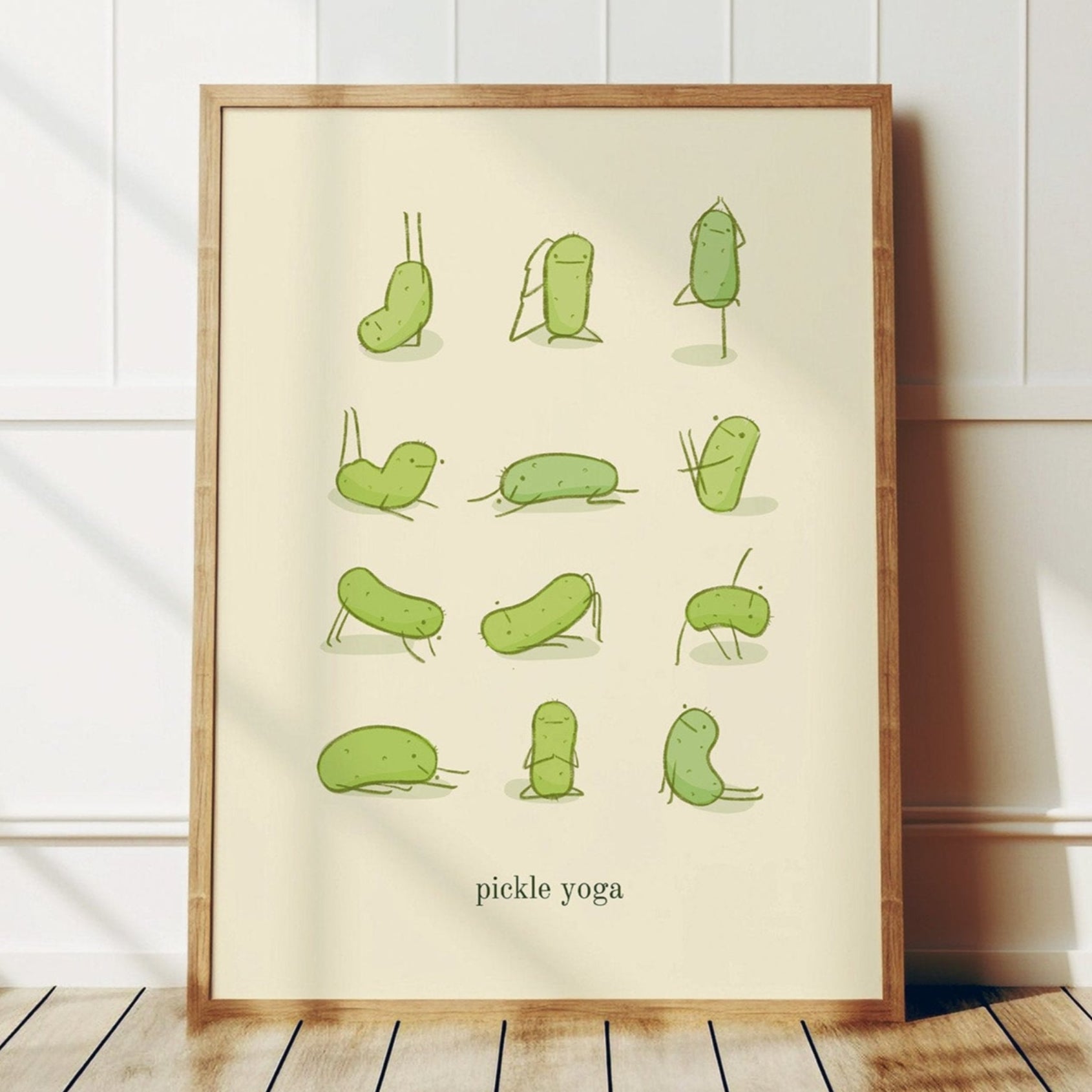 a funny pickle art print features a cute pickle in various yoga poses over text at the bottom that reads "pickle yoga" is displayed in a wooden frame that is leaning against a white wall