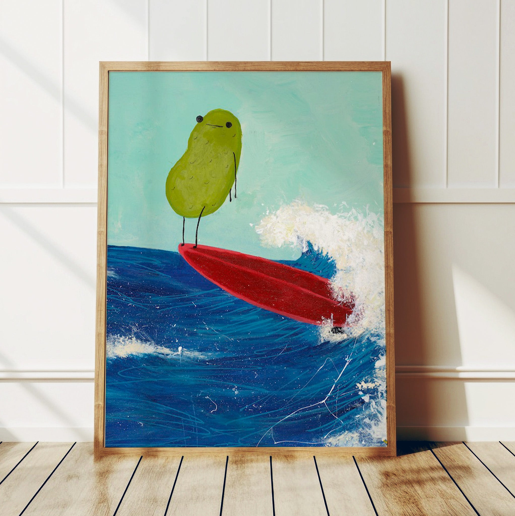 a pickle art print featuring a cute pickle riding a red surfboard on a wave is displayed in a wooden frame against a white wall in a brightly lit room
