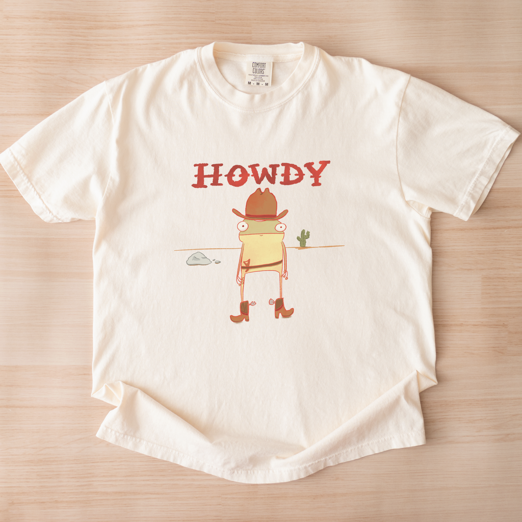 White t-shirt with a cartoon cowboy graphic and 'HOWDY' text on a wooden surface