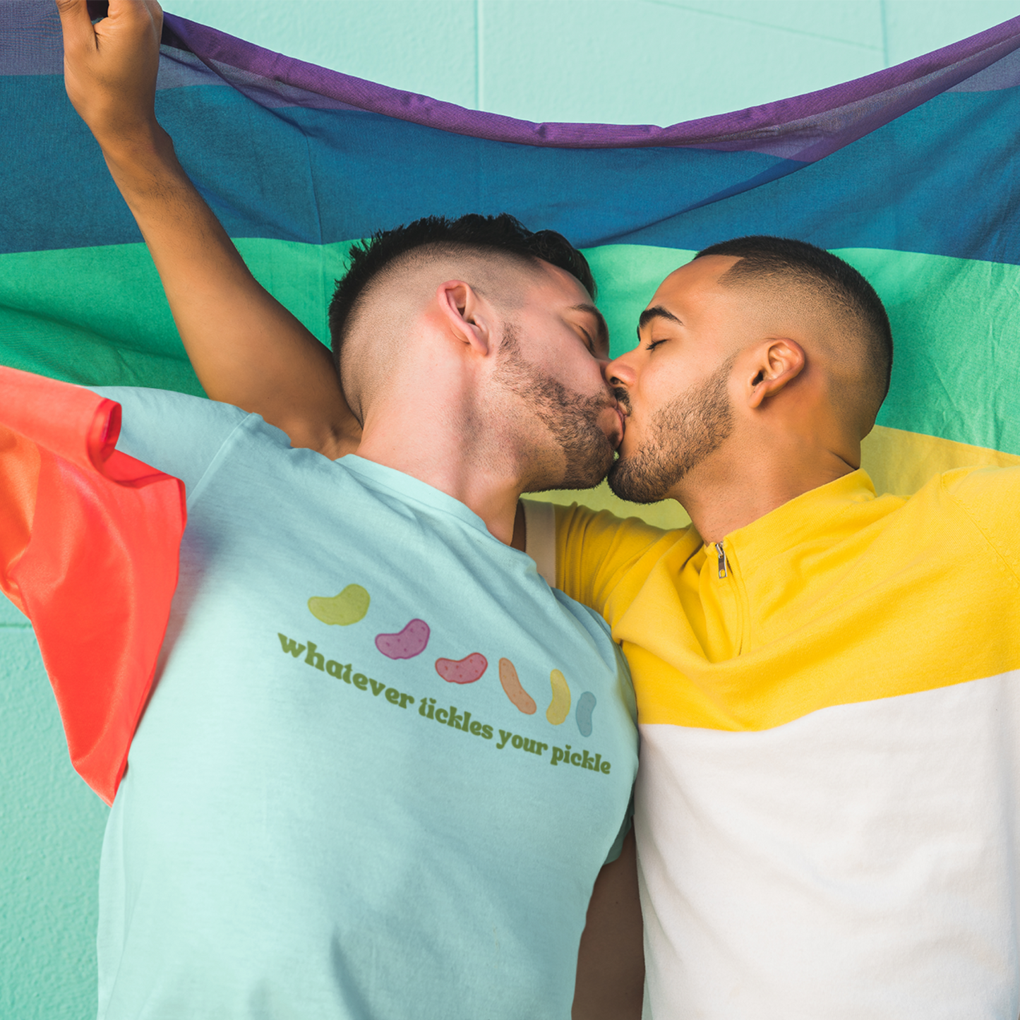 a gay couple kissing while holding the pride flag, with one person wearing a pickle themed pride shirt that says "whatever tickles your pickle"