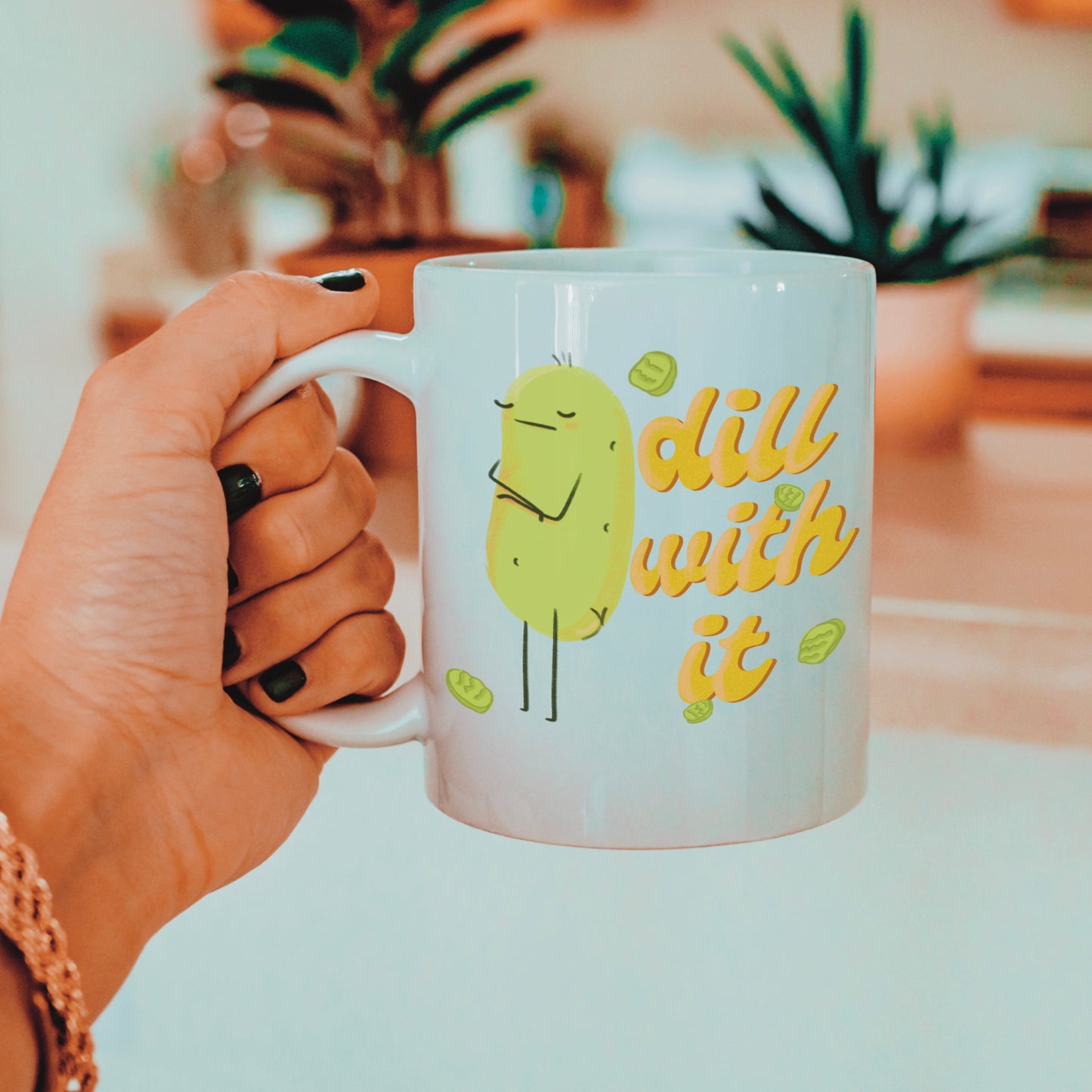 front view of a pickle mug featuring a cute illustration of a pickle character with its arms crossed next to funny text that says "dill with it" is held by a woman next to house plants