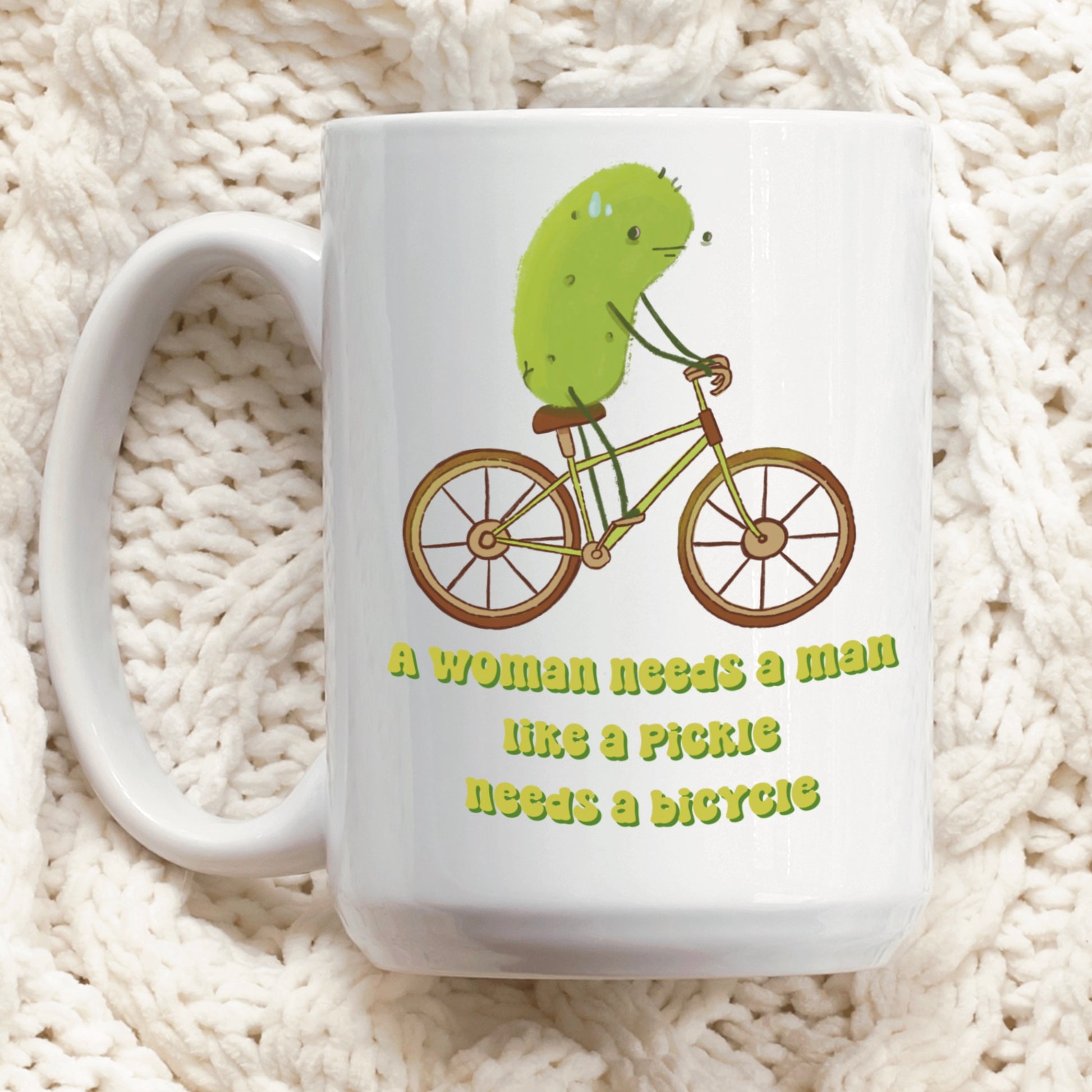 a feminist pickle mug featuring an illustration of a pickle on a bicycle and funny text that says "a woman needs a man like a pickle needs a bicycle" is displayed on a quilted table cloth