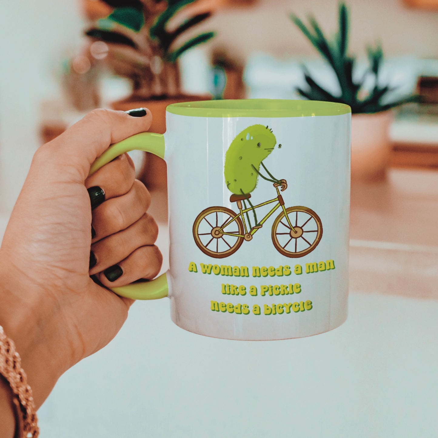 a feminist pickle mug featuring an illustration of a pickle on a bicycle and funny text that says "a woman needs a man like a pickle needs a bicycle" is held by a woman next to a house plant