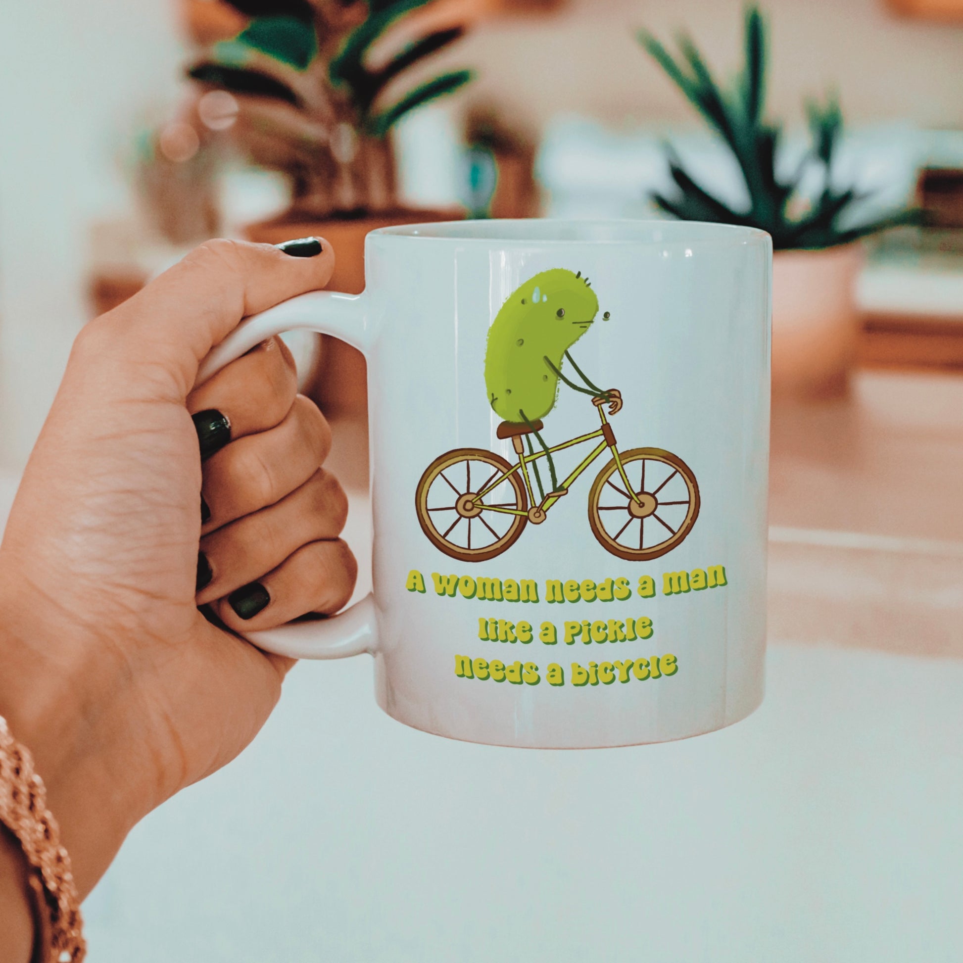 a feminist pickle mug featuring an illustration of a pickle on a bicycle and funny text that says "a woman needs a man like a pickle needs a bicycle" is held by a woman with gold jewelry