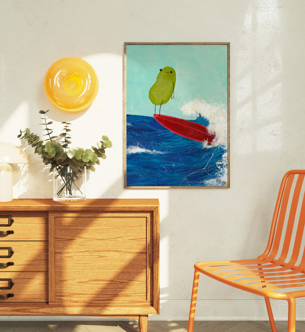 a pickle art print featuring a cute pickle riding a red surfboard on a wave is displayed in a wooden frame over a funky orange chair next to a house plant