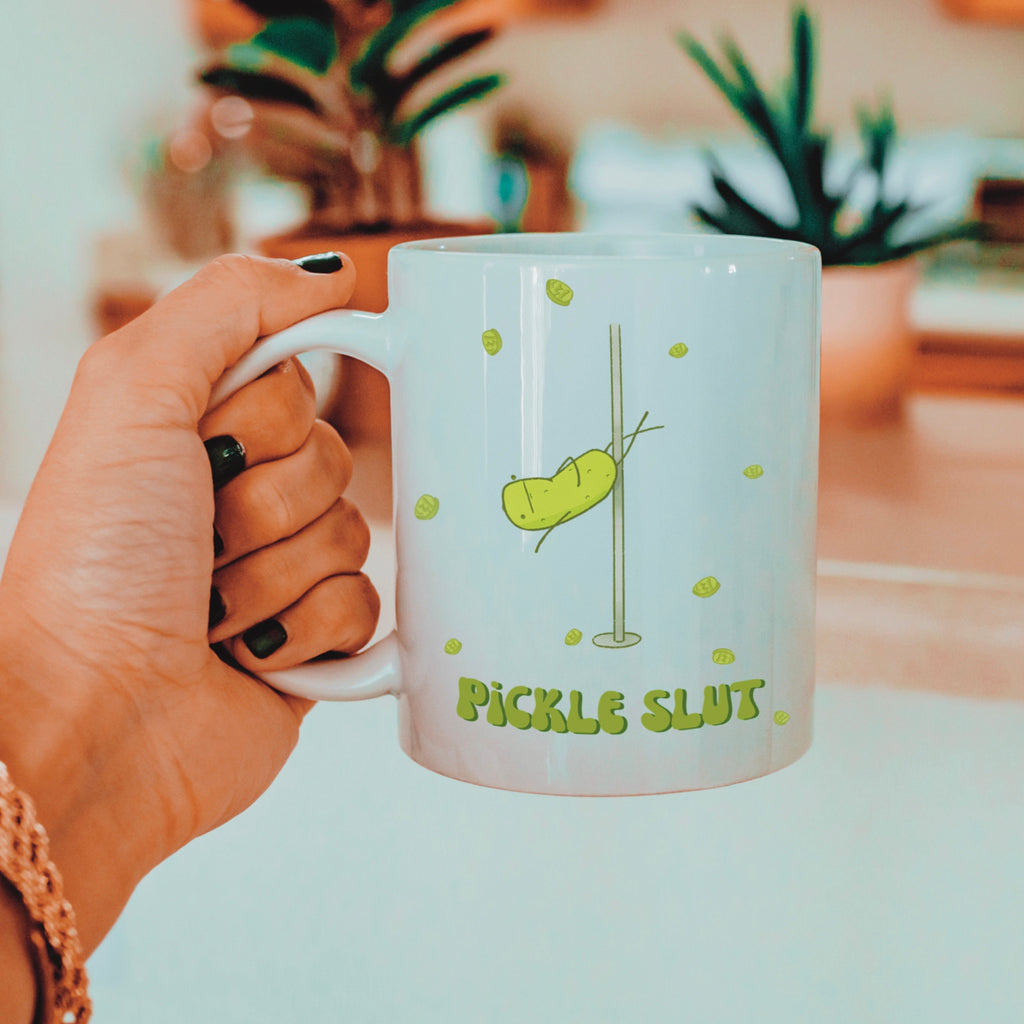 a pickle mug featuring a funny illustration of a cute pickle character pole dancing for pickle chips and text that says "pickle slut" is held by a woman wearing gold jewelry