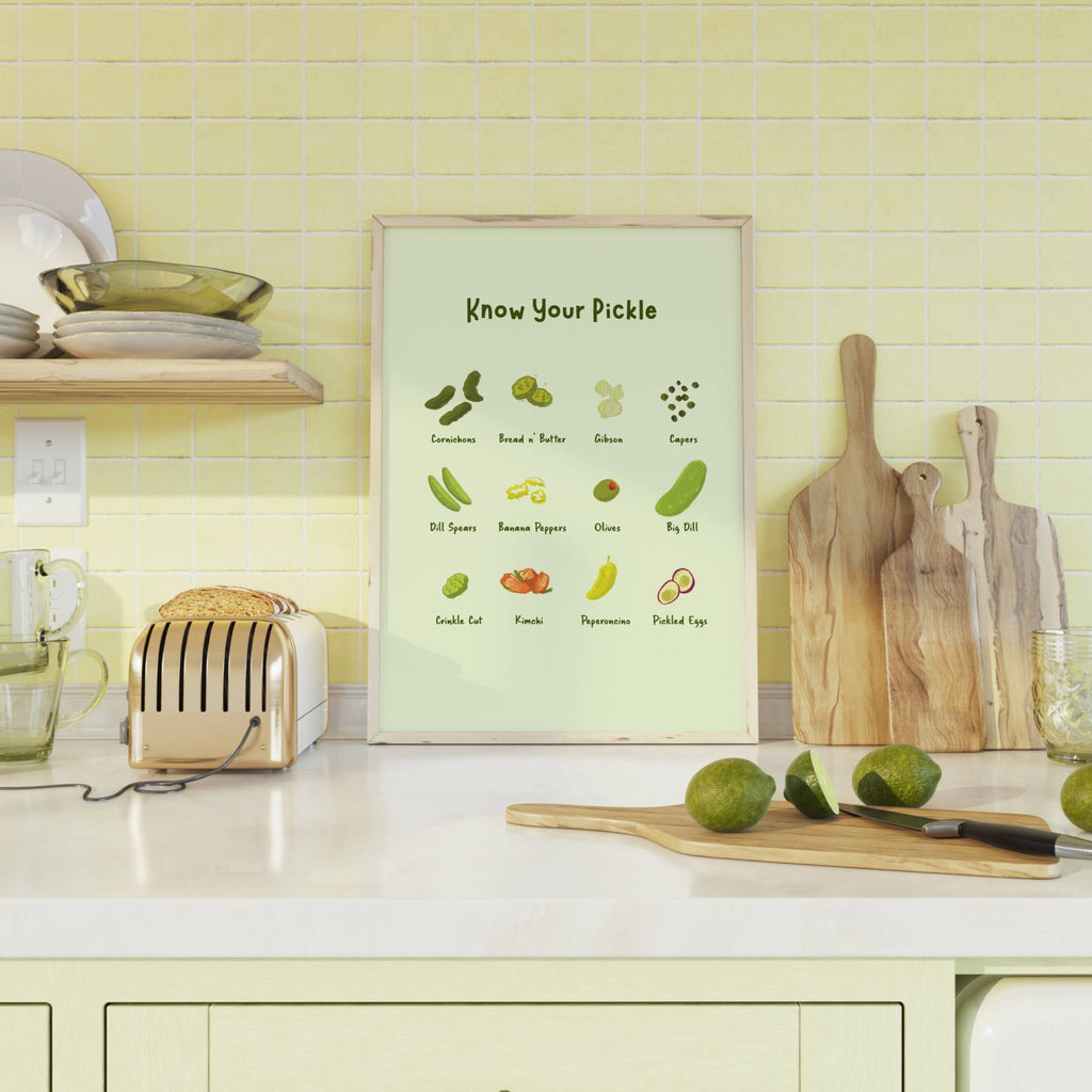 Pickle art featuring various pickles and their names is displaying in a wooden frame on a kitchen counter top next to cutting boards and a toaster