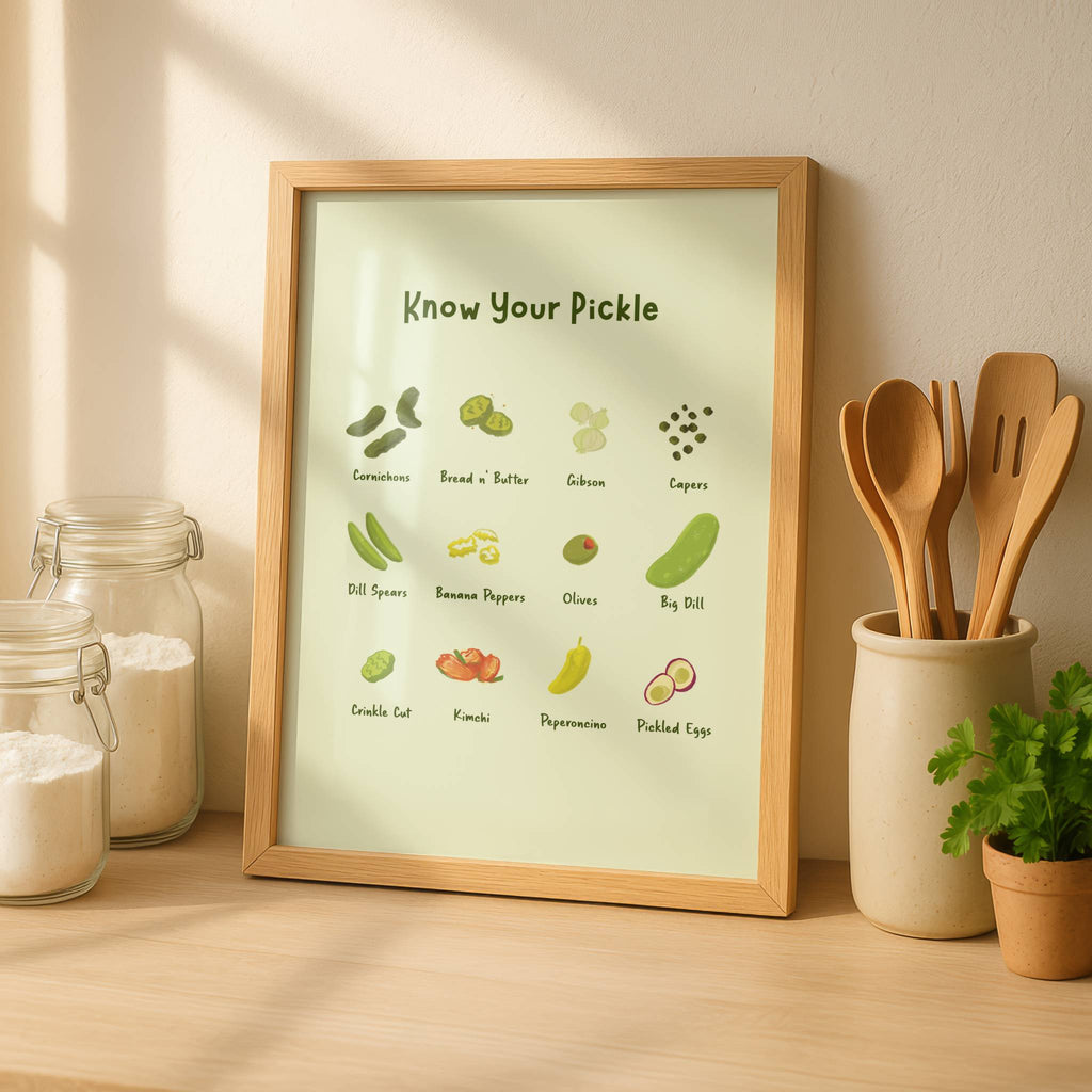 A pickle art print is displayed in a wooden frame next to some kitchen herbs and utensils