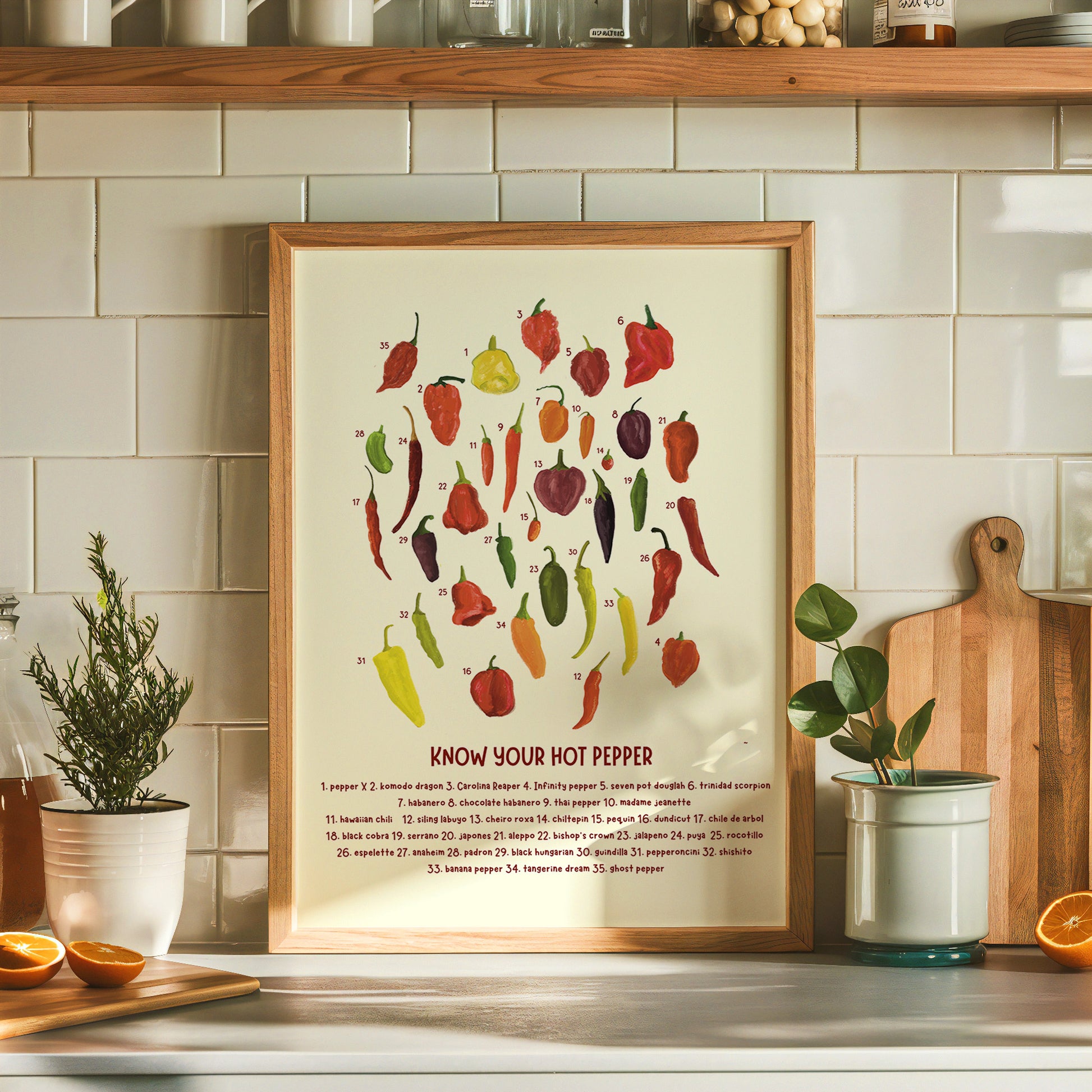 a hot chili pepper chart art print with an assortment of painted chilis are numbered over text that reads &quot;know your hot pepper&quot; with names of the numbered chili peppers is displayed in a wooden frame on a trendy kitchen countertop