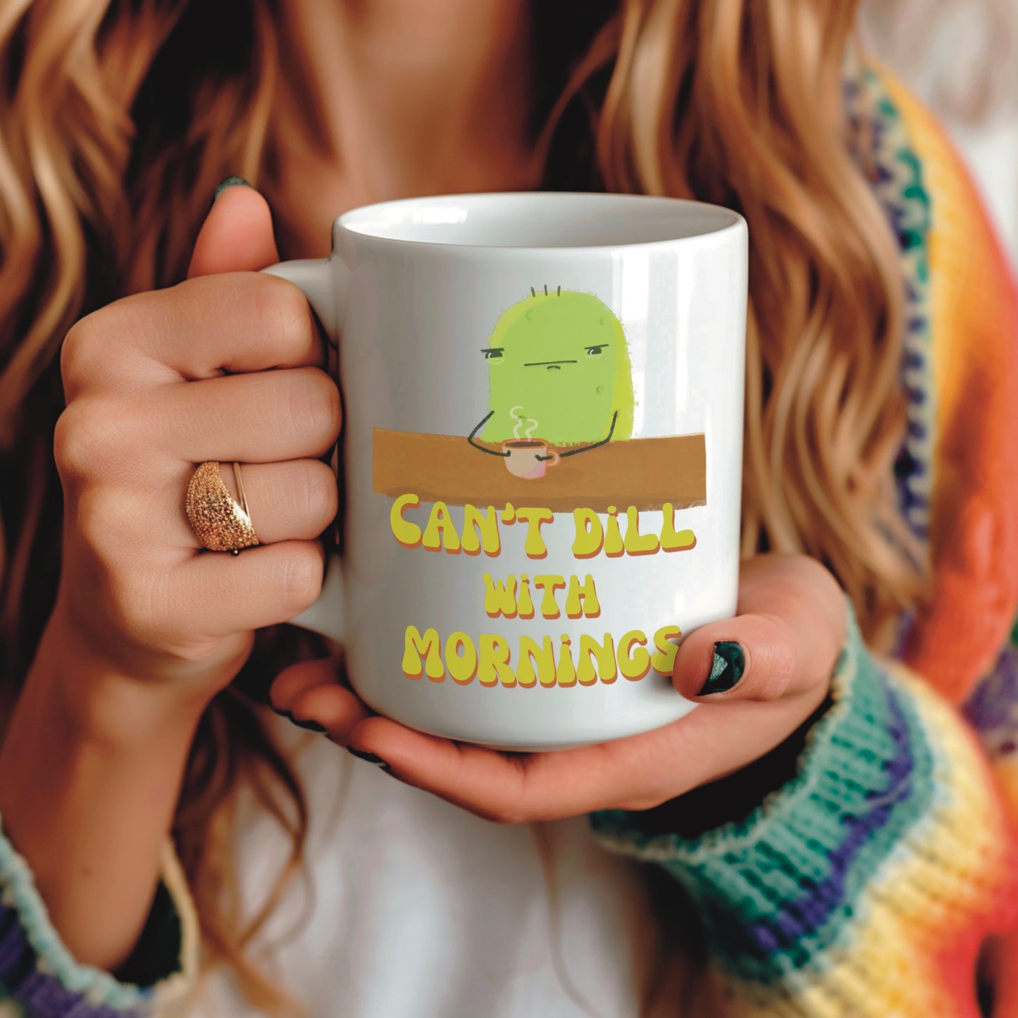 a pickle mug with a cute illustration of a pickle character drinking coffee over a punny quote that says "can't dill with mornings" is held by a woman in a rainbow sweater