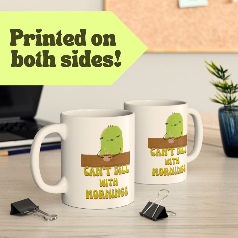 a pickle mug with a cute illustration of a pickle character drinking coffee over a punny quote that says "can't dill with mornings" is shown on a computer desk next to a house plant