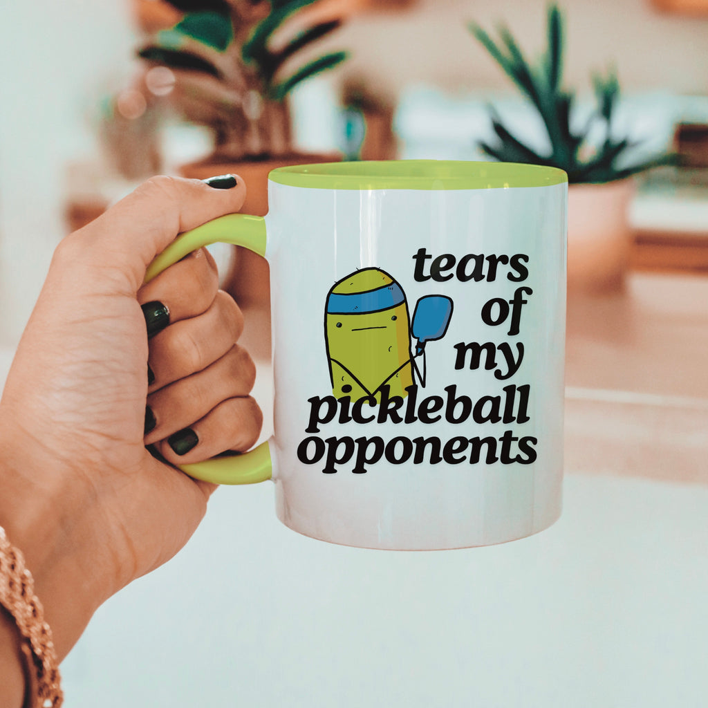 person holding a pickle ball mug with a funny quote that says "tears of my pickle ball opponents" with cute illustration of a pickle holding a blue pickle ball paddle