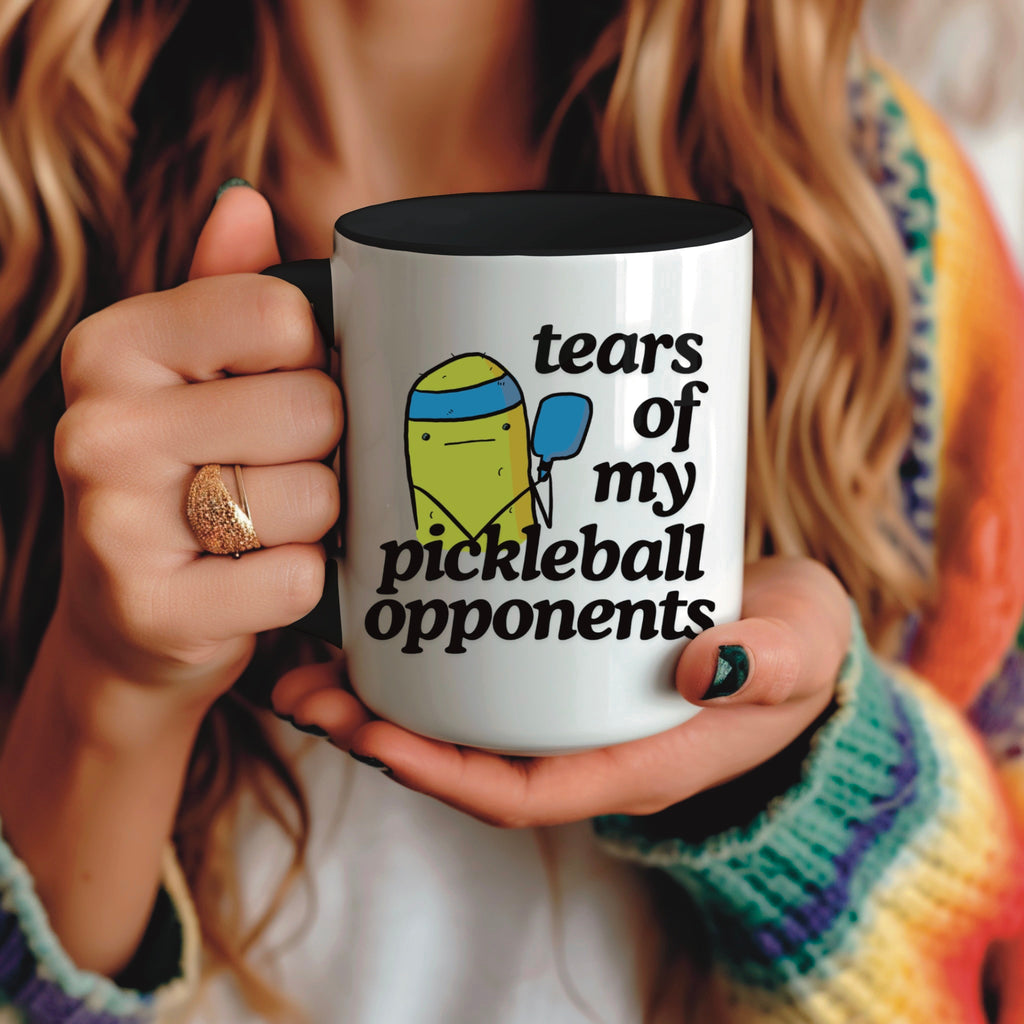 view of a woman holding a pickle ball mug with a funny quote that says "tears of my pickle ball opponents" with cute illustration of a pickle holding a blue pickle ball paddle