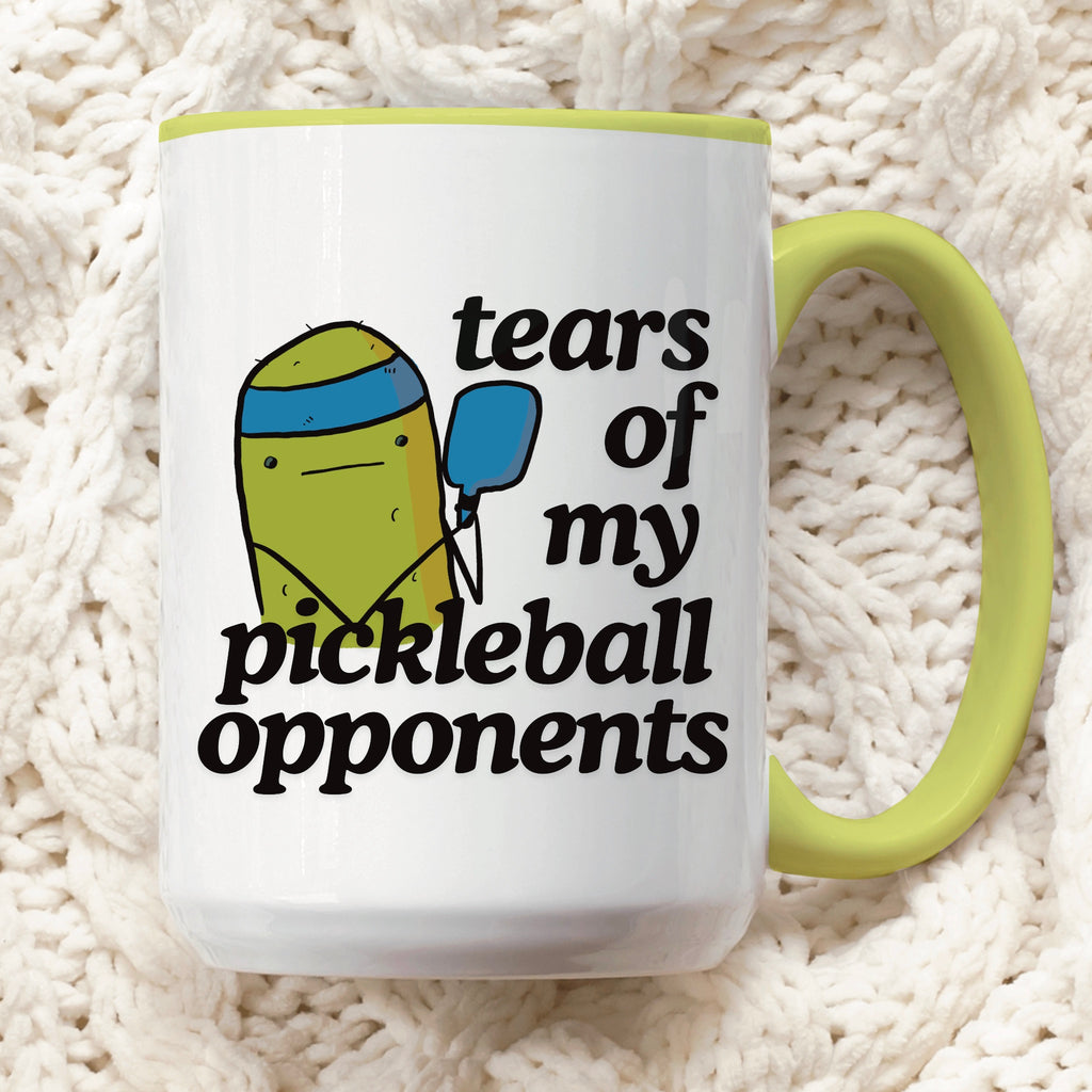 front view of a pickle ball mug with a funny quote that says "tears of my pickle ball opponents" with cute illustration of a pickle holding a blue pickle ball paddle