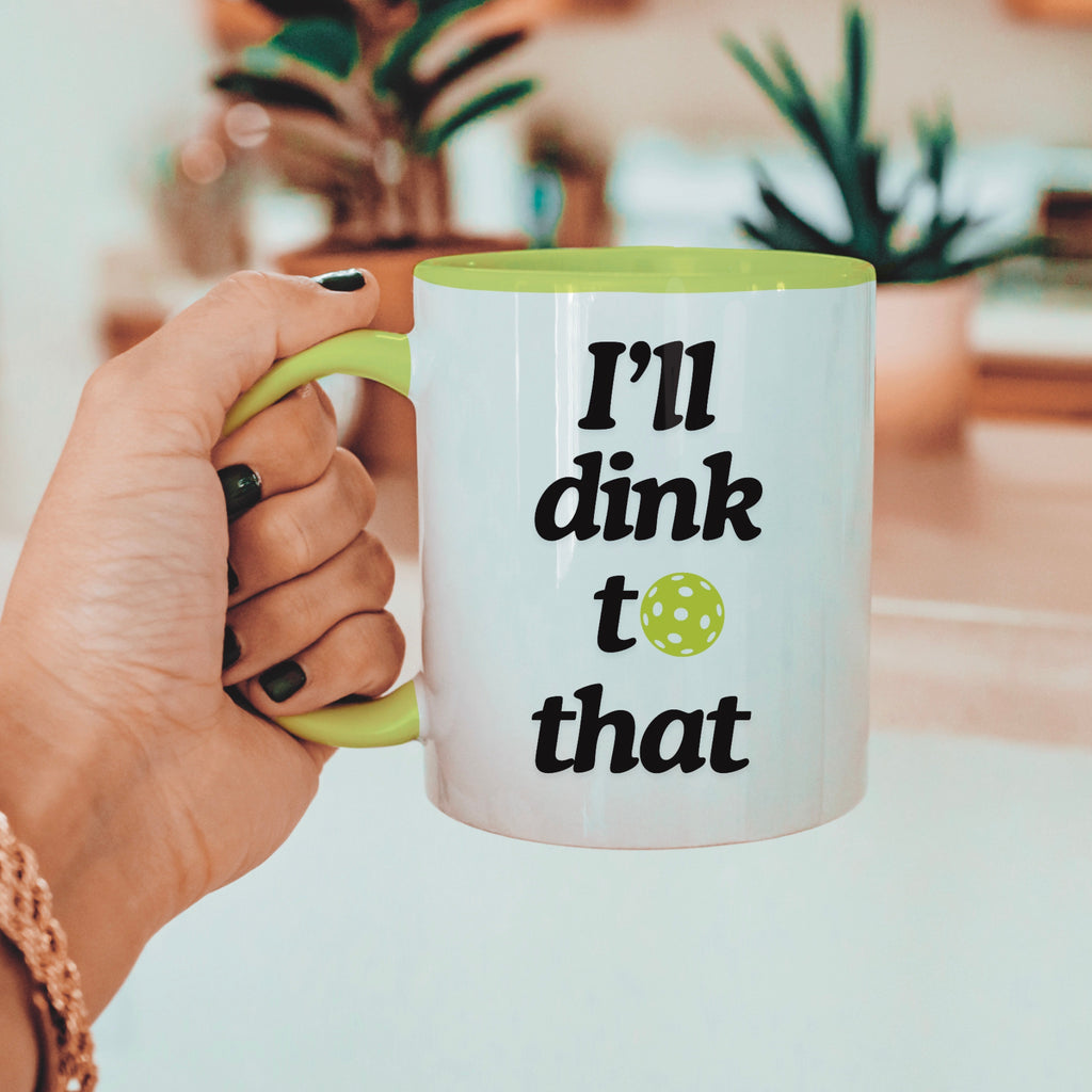 view of a hand holding a pickle ball mug with a funny quote that says "I'll dink to that" with illustration of a green pickle ball