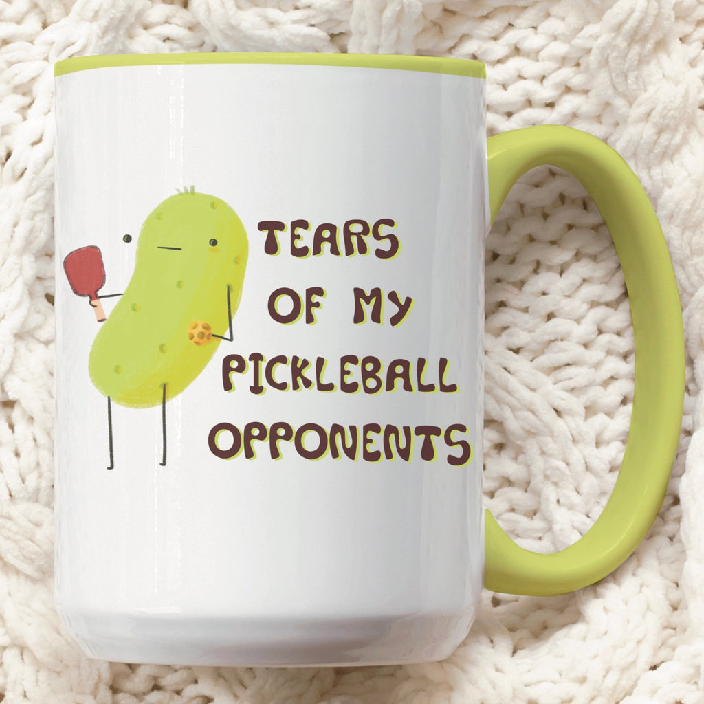front view of a pickleball mug with funny quote that says "tears of my pickleball opponents" next to illustration of a pickle holding a pickleball and paddle