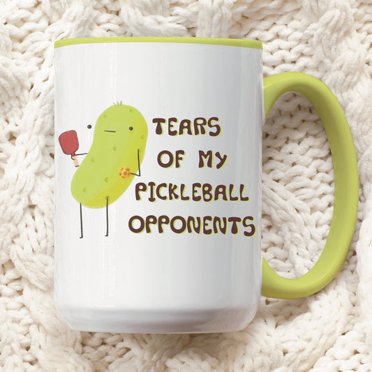 front view of a pickleball mug with funny quote that says "tears of my pickleball opponents" next to illustration of a pickle holding a pickleball and paddle