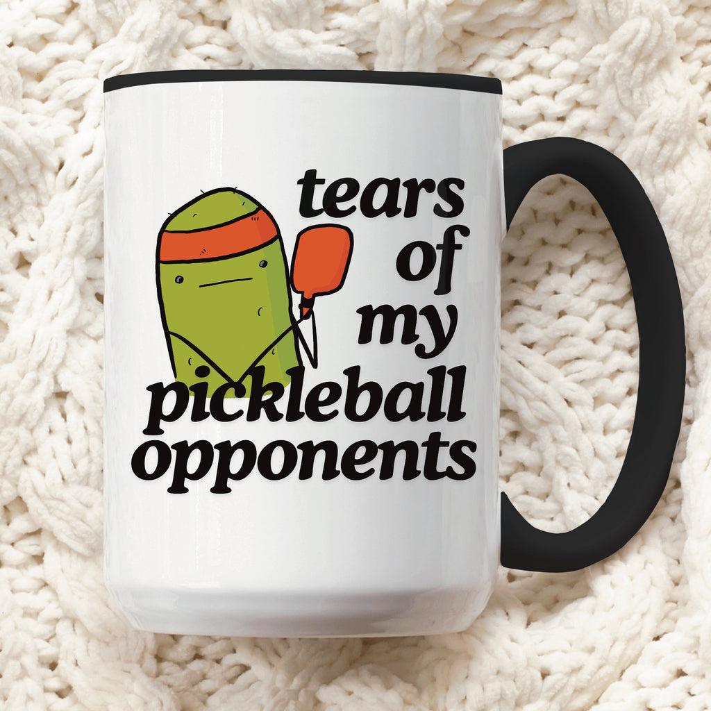 close up view of a pickle ball mug with a quote that says "tears of my pickle ball opponents" with cute illustration of a pickle holding a red pickleball paddle