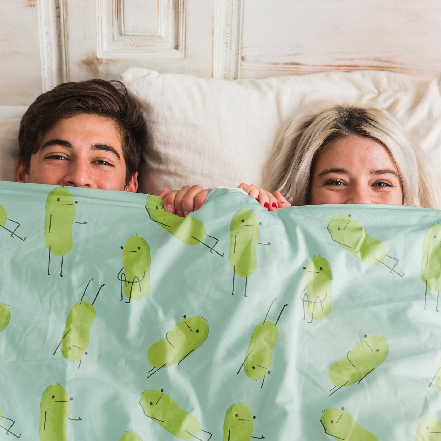 a pickle blanket that is light blue in color and has a print pattern of cute pickle characters in different positions is displayed on a bed with a smiling couple beneath it