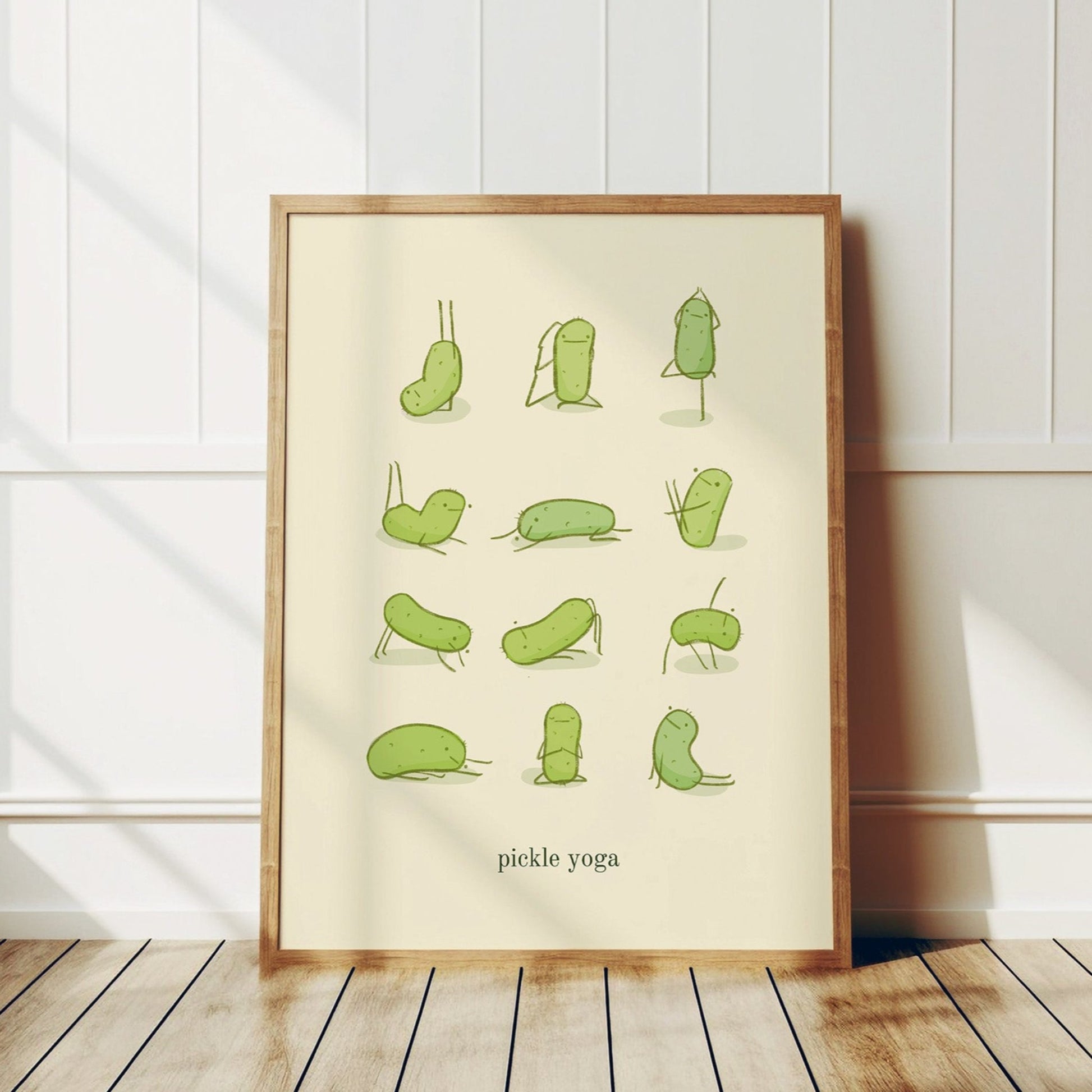 a funny pickle art print features a cute pickle in various yoga poses over text at the bottom that reads &quot;pickle yoga&quot; is displayed in a wooden frame that is leaning against a white wall