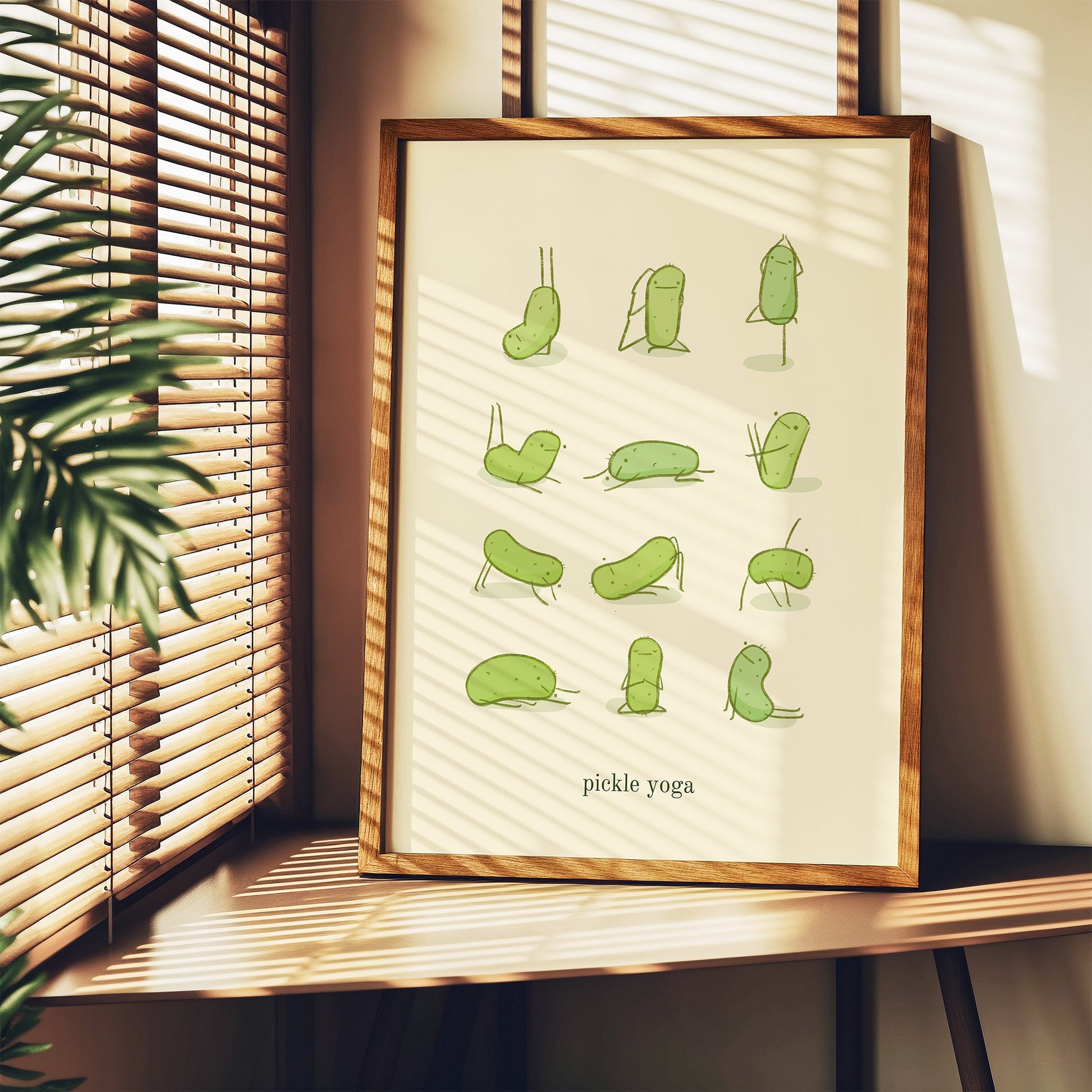 a funny pickle art print features a cute pickle in various yoga poses over text at the bottom that reads &quot;pickle yoga&quot; is displayed in a frame next to a window and a house plant